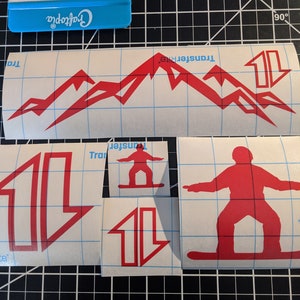 May include: Red vinyl sticker sheets with a mountain range, a snowboarder, and arrows. The sheets are on a gridded surface with a blue scraper tool.