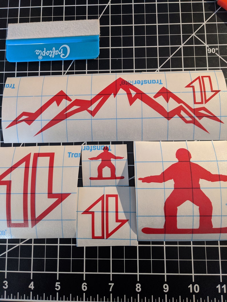 May include: Red vinyl sticker sheets with a mountain range, a snowboarder, and arrows. The sheets are on a gridded surface with a blue scraper tool.