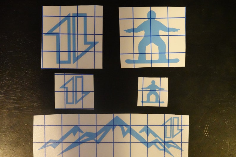 May include: Four blue vinyl stickers on a white gridded background. The stickers feature a mountain range, a snowboarder, and two different designs of an arrow pointing upwards.
