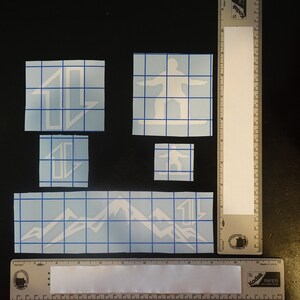 May include: Four white vinyl decals on a blue grid background. The decals include a mountain range, a snowboarder, and two different designs of a stylized letter "L".