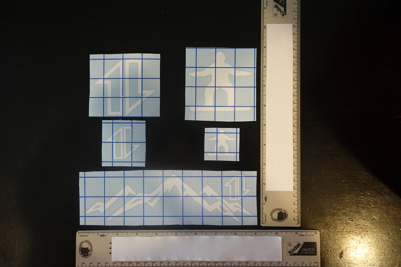 May include: Four white vinyl decals on a blue grid background. The decals include a mountain range, a snowboarder, and two different designs of a stylized letter "L".