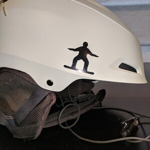 May include: White snowboard helmet with a black silhouette of a snowboarder sticker on the side. The helmet has a black mesh lining and a black strap.