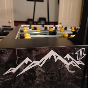 May include: Close-up of a black foosball table with a marble-like design. The table has a white mountain range logo with the word "11" on the side.