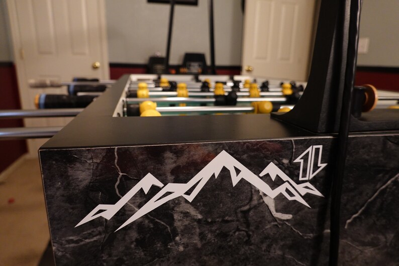 May include: Close-up of a black foosball table with a marble-like design. The table has a white mountain range logo with the word "11" on the side.