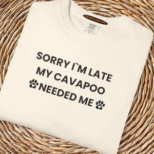 May include: Cream-colored t-shirt with the text "SORRY I'M LATE MY CAVAPOO NEEDED ME" in black. The shirt features paw print graphics. The tag reads "Comfort Colors 1717".