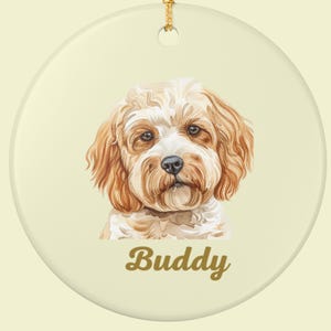 May include: Round ceramic ornament featuring a watercolor portrait of a dog with tan and white fur. The dog's name, "Buddy," is written in a cursive font below the portrait. A gold-colored string is attached for hanging.