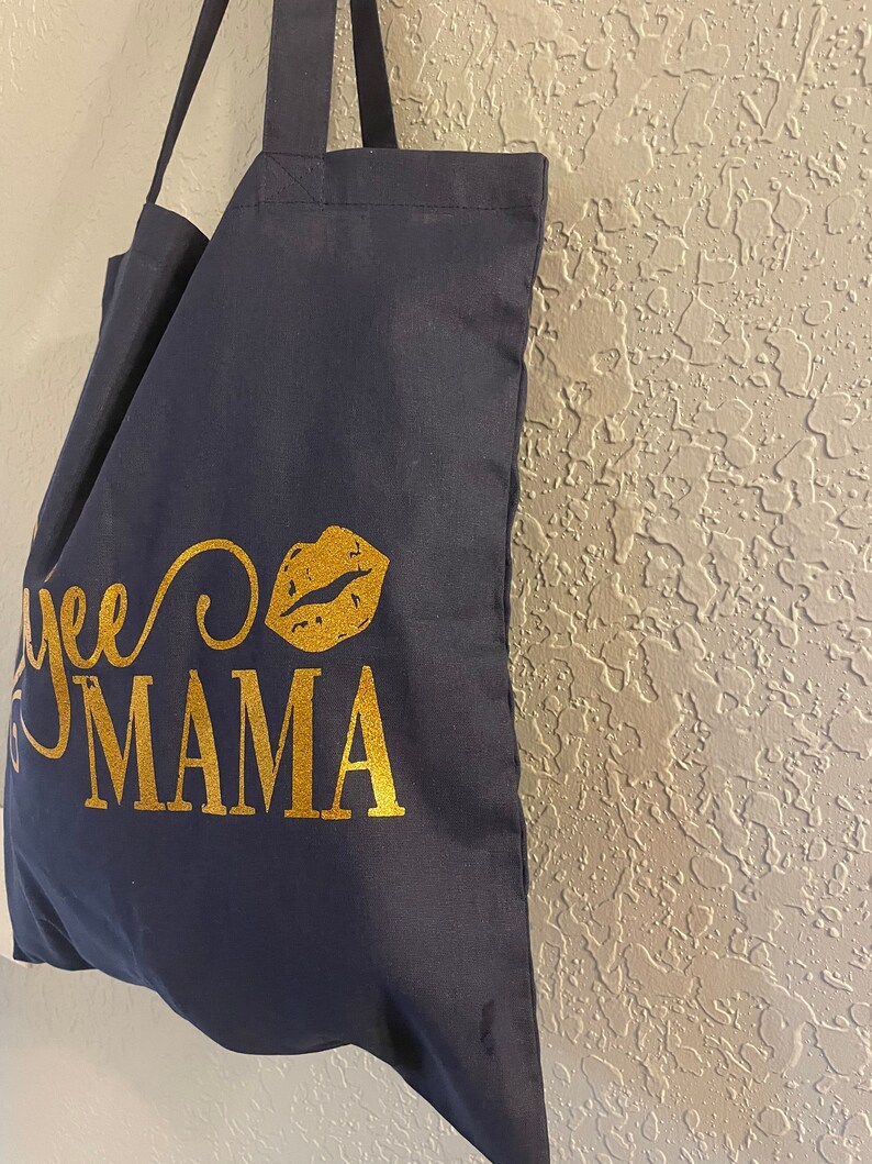 May include: A navy blue tote bag with gold lettering that says "Yee to Mama" with a gold glitter lip print.