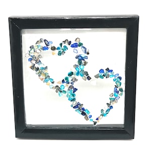 May include: A framed artwork featuring two overlapping hearts. The hearts are composed of small, irregularly shaped stones in shades of blue, teal, and gray. The frame is black, and the background is white, creating a striking contrast.
