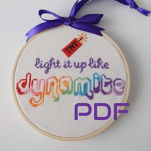 May include: A white embroidery hoop with a rainbow colored word "dynamite" and the text "light it up like" above it. A small red and yellow dynamite stick is above the word "dynamite". The hoop has a purple ribbon tied around it. The text "PDF" is in purple below the hoop.
