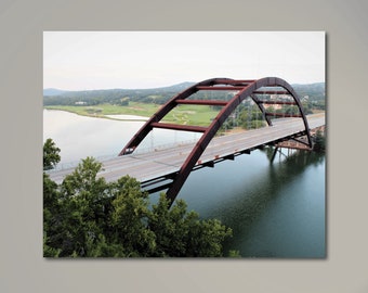 May include: A red steel bridge arches over a body of water with green trees and a golf course in the background.