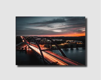 May include: A long exposure photograph of a bridge with traffic lights streaking across the roadway. The bridge is lit up with warm yellow lights and the sky is a vibrant orange and pink.