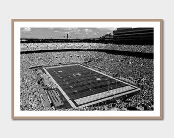 May include: A black and white photograph of a packed football stadium with a football field in the center. The field is marked with yard lines and a large 'H' at midfield.