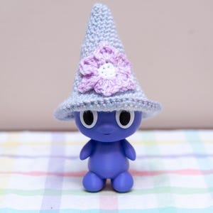 May include: A blue figurine wearing a light blue crocheted hat with a purple flower. The figurine has large black eyes and small arms and legs. The background includes a decorative figure and a patterned surface.