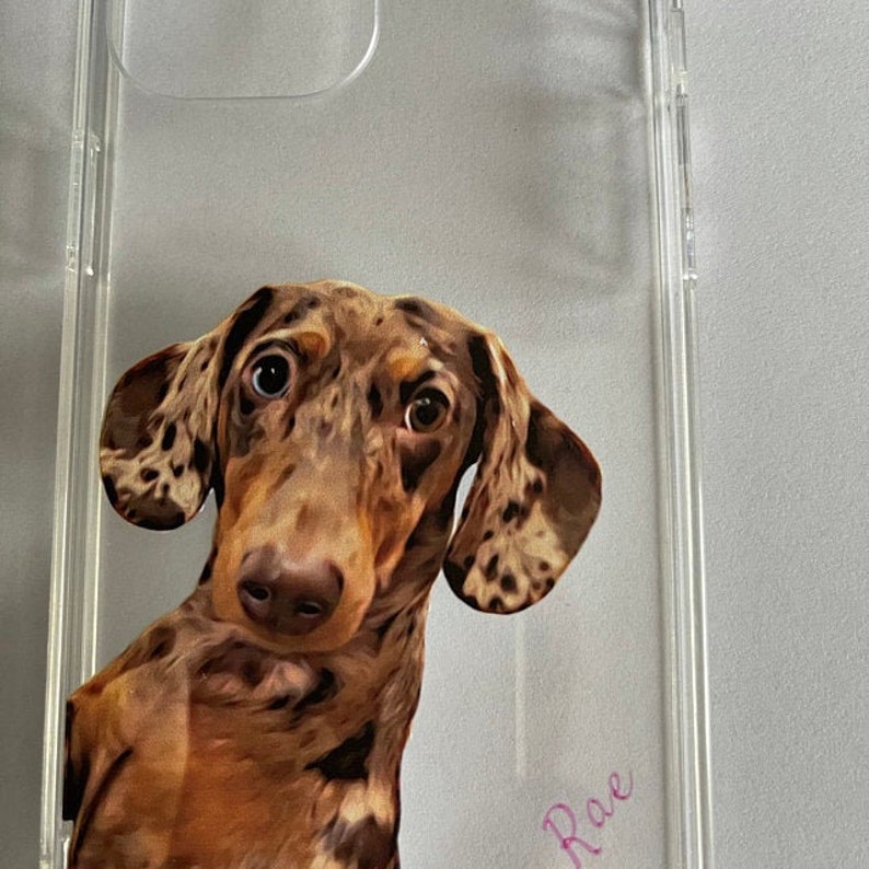Może przedstawiać: Przezroczyste etui na telefon z ilustracją brązowego i czarnego psa rasy dachshund. Imię psa, "Rae", jest napisane na etui r&oacute;żowym kolorem.