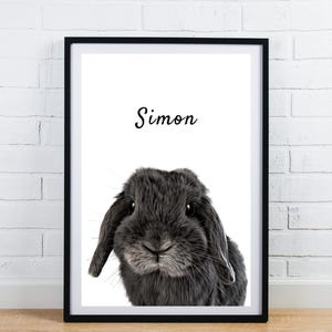 May include: Framed print featuring a close-up of a gray rabbit with floppy ears. The name "Simon" is written in cursive above the rabbit. The print is in a black frame and is displayed against a white brick wall.