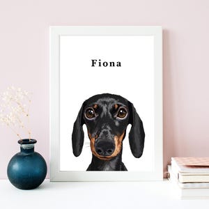 May include: A framed art print featuring a black and tan dachshund dog with the name "Fiona" above the dog's head. The print is in a white frame, and the background is white. A small, dark blue vase with white flowers is on the left.
