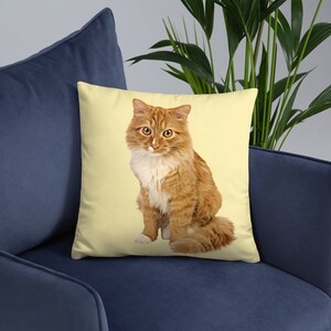 Puede incluir: Coj&iacute;n cuadrado con un gato jengibre con marcas blancas, sobre un fondo amarillo p&aacute;lido. El gato est&aacute; sentado, con la cola enroscada. El coj&iacute;n est&aacute; colocado en un sof&aacute; azul.
