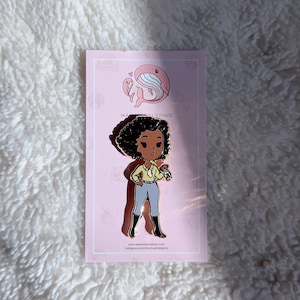 Pode incluir: Um alfinete com um personagem de desenho animado com pele escura, cabelo encaracolado e uma camisa amarela, calças azuis e botas pretas. O alfinete está em um cartão rosa com uma ilustração de baleia e o texto "Mama Hale Pins".