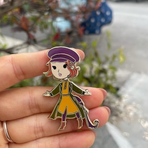 Puede incluir: Un pin de esmalte dorado con un personaje de dibujos animados que lleva un sombrero morado, un abrigo verde y un vestido amarillo. El personaje tiene el pelo rojo y lleva un bolso verde.