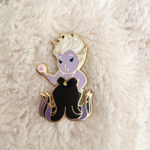 May include: A purple and white enamel pin of Ursula from The Little Mermaid. She is holding a pink seashell in her right hand and has a black dress with a gold necklace. Her tail is purple and black with gold accents.
