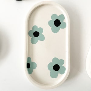 May include: A white oval tray with a raised rim, decorated with four light blue flowers with black centers. The tray is perfect for holding jewelry, keys, or other small items.