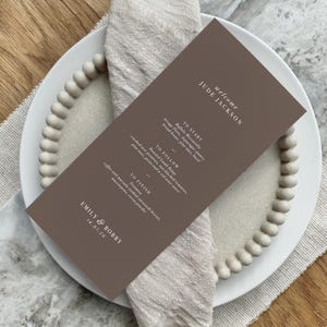 以下が含まれることがあります： 白い文字が入った茶色のメニューカード。ビーズの縁が付いた白いお皿とベージュのリネンのナプキンに置かれています。メニューには3コースの食事内容が記載されています。メニューはエミリー＆ボビーの結婚式用です。