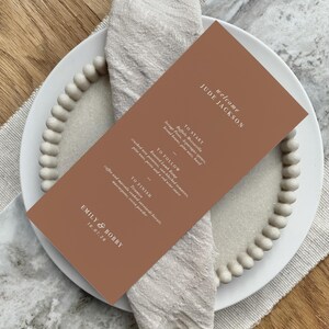 Könnte beinhalten: Eine terrakottafarbene Hochzeitsmenükarte mit weißem Text, die auf einem weißen Teller mit Perlenrand und einer Leinen-Serviette liegt. Das Menü enthält die Namen "Jude Jackson" und "Emily & Bobby" mit einem Datum.