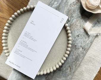 May include: A white menu card with the text "the MENU" and a list of dishes, placed on a beige beaded plate. The plate sits on a marble-patterned surface with a beige linen runner. The menu includes the names of dishes and the names Emilia & MARK.