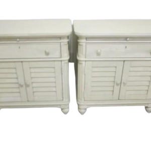May include: A pair of white wooden nightstands with a single drawer and two louvered doors. Each nightstand has a simple design with turned legs.