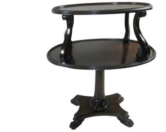 May include: A dark brown wooden two-tiered table with an oval top and a pedestal base. The table has ornate scrollwork details on the legs and a decorative beaded edge on the top tier.