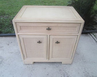 May include: A white wicker end table with a single drawer and two doors. The table has brass hardware and a woven top.