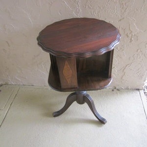 May include: A dark wood, round, revolving bookcase table with a scalloped edge and a diamond-shaped inlay. The table has three legs and is on a light beige floor.