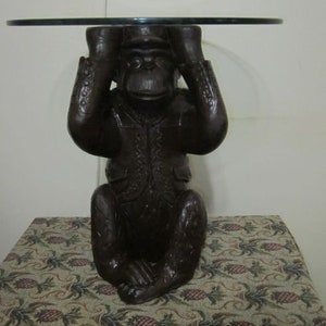 May include: A dark brown wooden monkey sculpture with a glass top table. The monkey is covering its eyes with its hands.