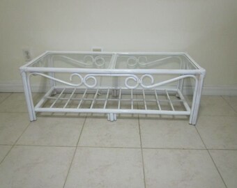 May include: White wicker coffee table with a glass top and a lower shelf. The table has a decorative scroll design on the sides.
