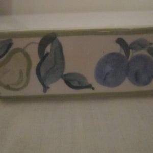 May include: Rectangular ceramic dish with a cream-colored base. The dish is decorated with hand-painted green pears and blue plums with green leaves. The rim of the dish is outlined with a green border.