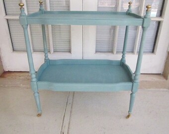 May include: A two-tiered serving cart with a light blue finish and gold accents. The cart has a rectangular top and bottom shelf and four legs with decorative details. The cart is on wheels.