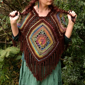 May include: A handmade crochet poncho in shades of brown, blue, green, and yellow. The poncho features a geometric design with a central square motif and fringe along the bottom edge. The garment is worn over a green top and skirt.