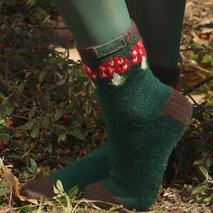 May include: Green knitted socks with a brown heel and toe, adorned with a mushroom design in red and white on the cuff. The socks are worn with green tights, set against a backdrop of foliage with red berries.