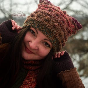 Può includere: Un cappello di lana marrone con un design strutturato e a pompon, abbinato a guanti marroni e una sciarpa. Il cappello passa dal marrone al rosso e al viola. La sciarpa ha strisce rosse, arancioni e verdi. Il logo Forest LaNa è visibile.