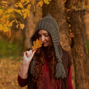 May include: A woman wearing a grey knitted hooded hat with tassels, a burgundy shirt, and a brown vest. She is holding a brown leaf in her hand and is standing in a forest with autumn leaves on the ground.
