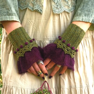Può includere: Guanti senza dita verde oliva e viola con bordo all'uncinetto e piccoli pompon viola. I guanti hanno un polsino a coste e un polso viola svasato. L'immagine mostra i guanti indossati.