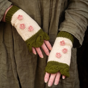Puede incluir: Guantes sin dedos color crema con tres aplicaciones de flores rosas. Los puños y los bordes son de color verde oscuro con un diseño de hojas festoneadas. Los guantes están tejidos y tienen una estética caprichosa de cuento de hadas.