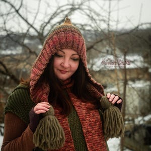 Pode incluir: Um conjunto de gorro e cachecol de malha em tons de castanho, vermelho e verde. O gorro tem um topo pontiagudo e o cachecol apresenta grandes borlas. O conjunto foi concebido para aquecer e dar estilo, ideal para o frio.
