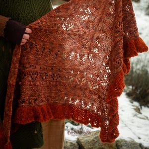 May include: A rust-colored knitted shawl with intricate openwork and a ruffled edge. The shawl is held up, showcasing its delicate design and texture. The color is a warm, earthy tone, perfect for fall or winter wear.