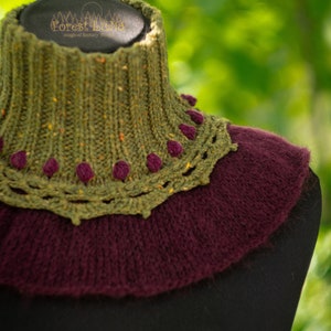 May include: A knitted cowl in olive green and deep burgundy, displayed on a black mannequin. The cowl features a textured, ribbed design with a decorative scalloped edge and small, dark purple accents. The background is a soft, blurred green.