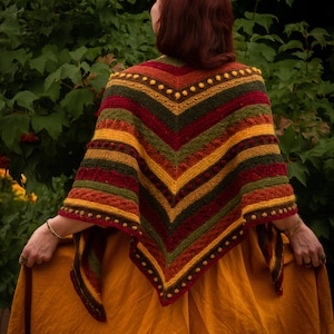 Może przedstawiać: Kobieta w kolorowym, paskowanym szalu na szydełku z fakturowanym wzorem i brązowymi pomponami. Szal jest zarzucony na długą, musztardowożółtą sukienkę.