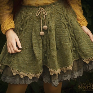 Könnte beinhalten: Olivgrüner Strickrock mit Zopfmuster, Kordelzug in der Taille und dekorativen Bommeln. Der Rock hat ein mehrschichtiges Design mit Spitzenbesatz und eine fließende Silhouette, ideal für einen verspielten, böhmischen Look.