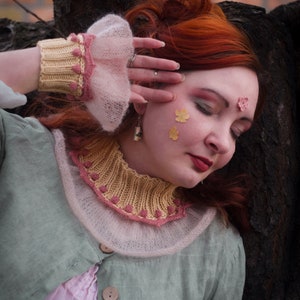Puede incluir: Una mujer pelirroja con una chaqueta verde claro y un vestido rosa, con un cuello y puños tejidos en amarillo y rosa. Decoraciones con forma de flor adornan su rostro. La imagen tiene un ambiente artístico.