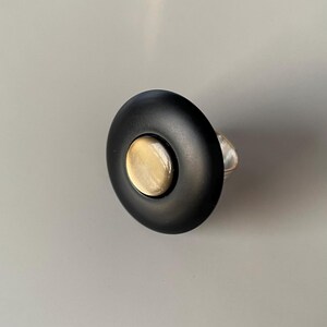 May include: A black ring with a circular design. The ring has a large, flat, black surface with a smaller, gold-coloured circle in the centre. The band of the ring is silver-coloured.