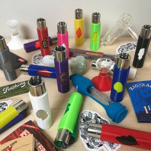 May include: A collection of colorful lighters with various designs and patterns. The lighters are arranged on a wooden surface with other smoking accessories, including a glass pipe, a grinder, and rolling papers. The lighters are made of metal and have a variety of colors, including red, blue, green, yellow, and purple.